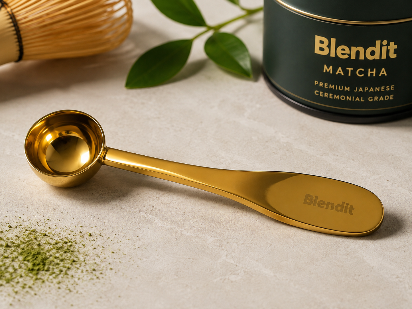 A polished gold Blendit measuring spoon with a long handle lies on a light stone surface, with a dark green Blendit Matcha tin, green leaves, and a bamboo whisk in the background.