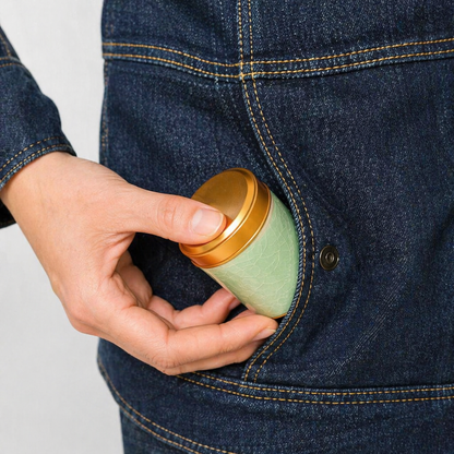 Person putting a green and gold container into their blue denim jacket pocket.